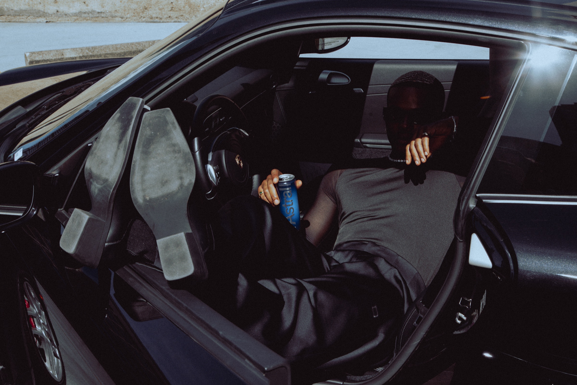 Model in black leaning back in a Porsche driver's seat, boots up on the open door, holding a can of Astral