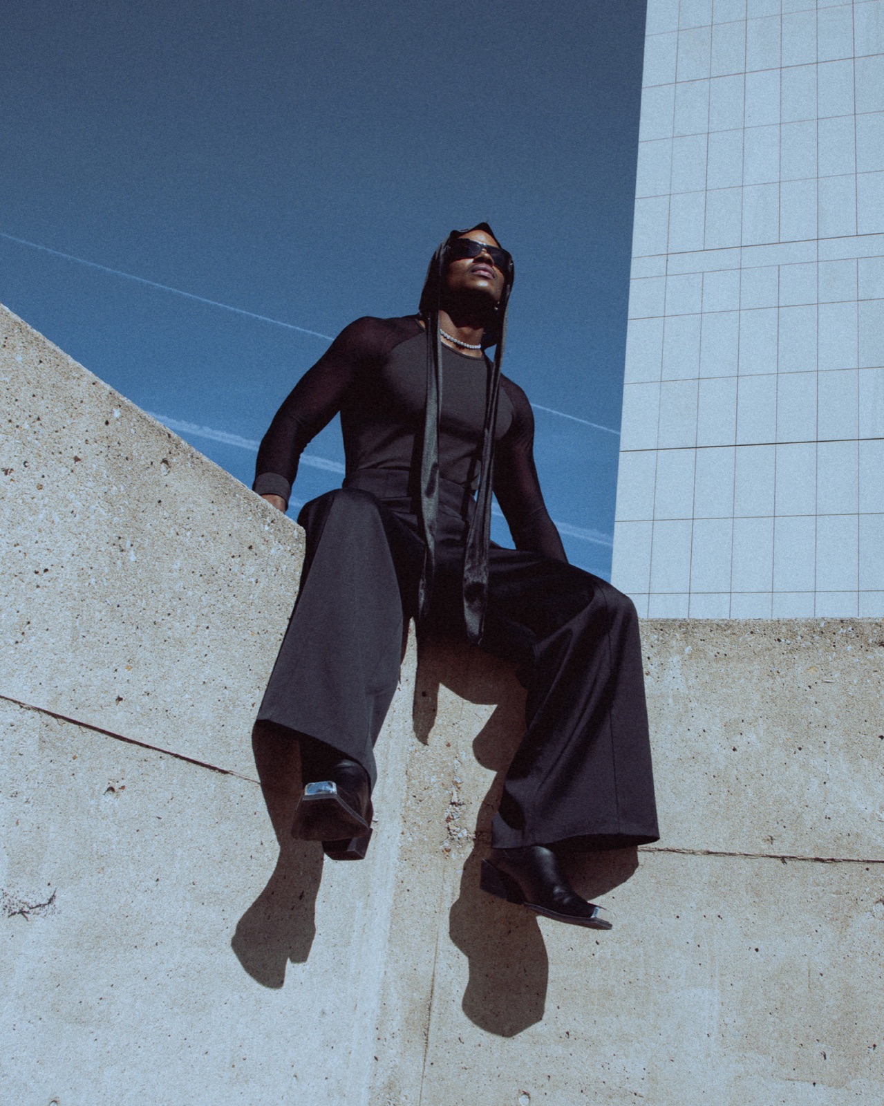 Model in black sitting on a concrete wall, fashion editorial
